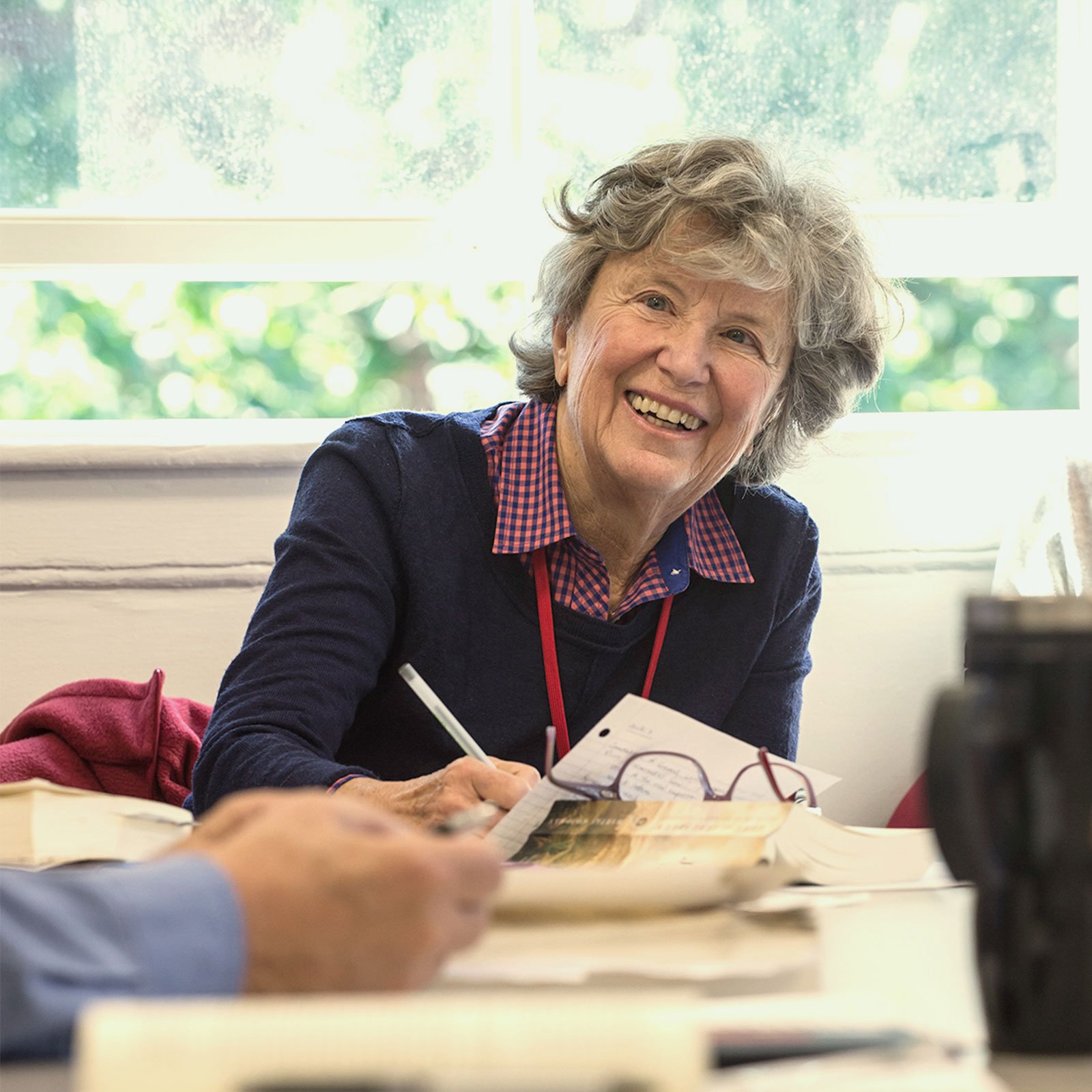 A participant of Harvard's Institute for Learning In Retirement sits at a table, smiling and looking at someone off camera.